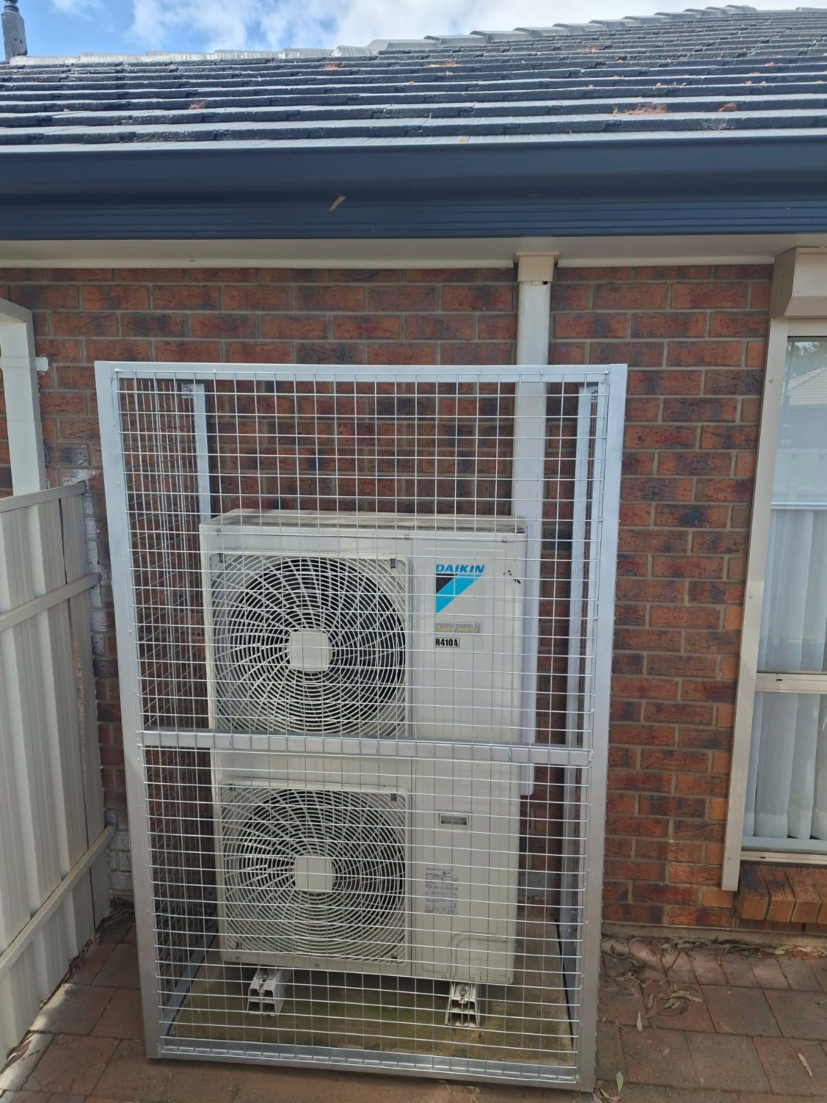 Galvanised steel mesh security cage built around an air-conditioner outdoor unit against a brick house wall